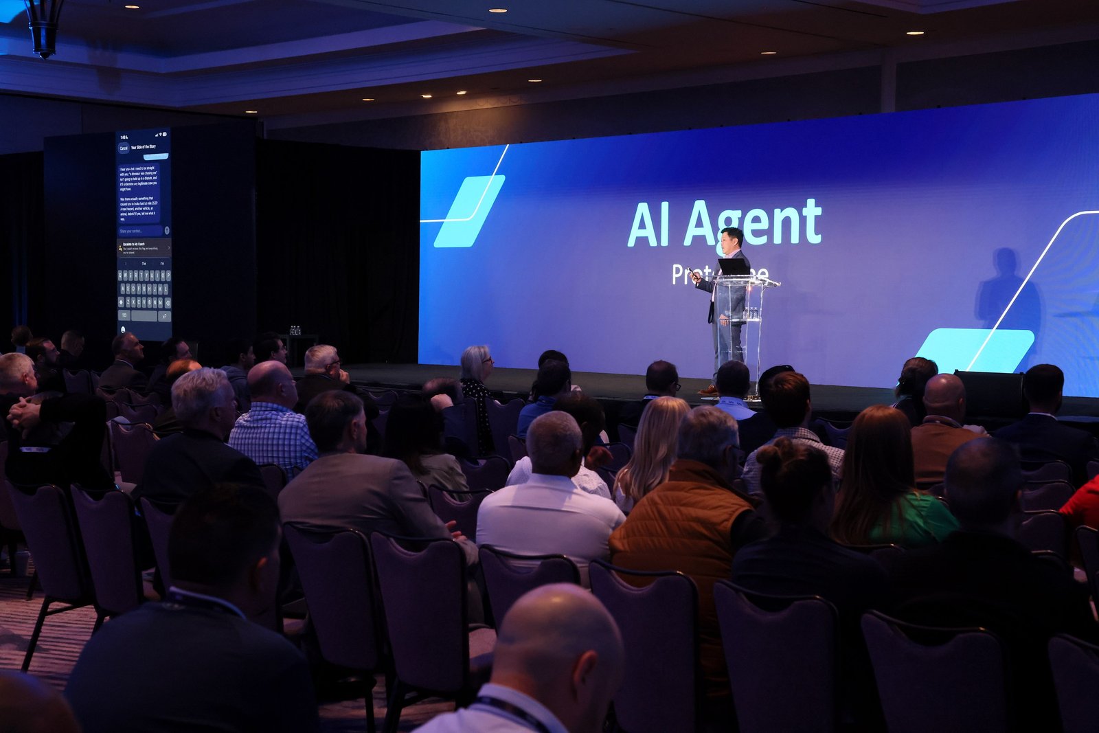 A presenter onstage beside a projected mobile application and the title AI Agent Prototypes, with an audience in the foreground.